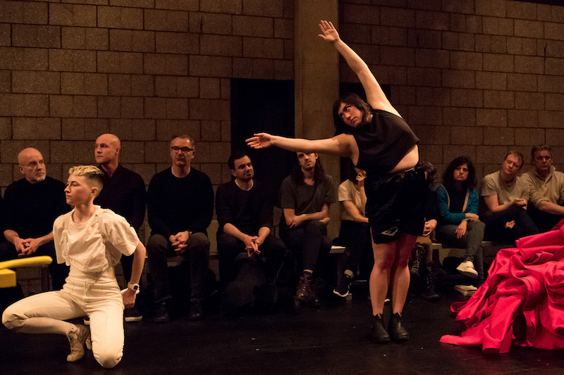 Jen Rosenblit in black boxing shorts and top reaches her arms over her head to the side. A performer in all white crouches to the right of her.